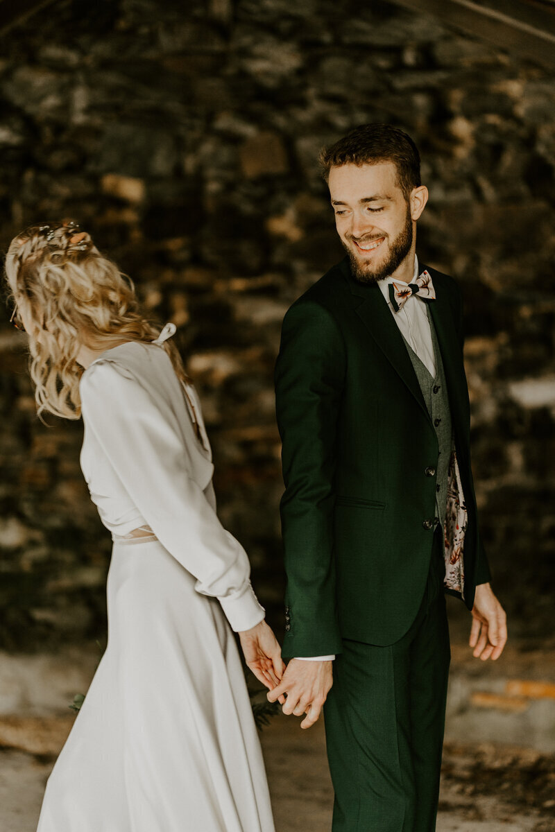 Mariée tournant le dos au marié, souriant, lui tenant la main dans un écrin de verdure. Immortalisés par Laura Termeau, photographe professionnelle en Vendée.