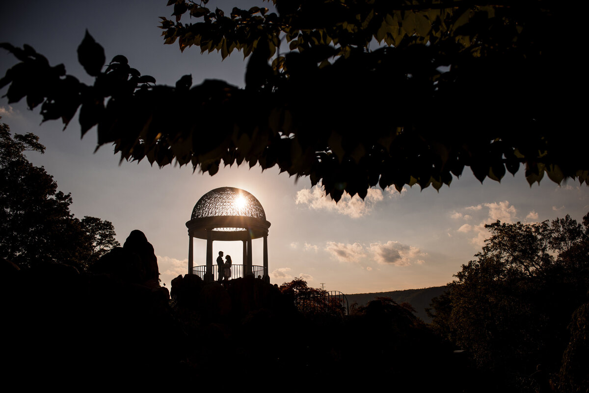 Silhouetted couple stands under a decorative gazebo at sunset, surrounded by trees and an expansive sky at an Untermeyer Gardens sunset engagement photo shoot