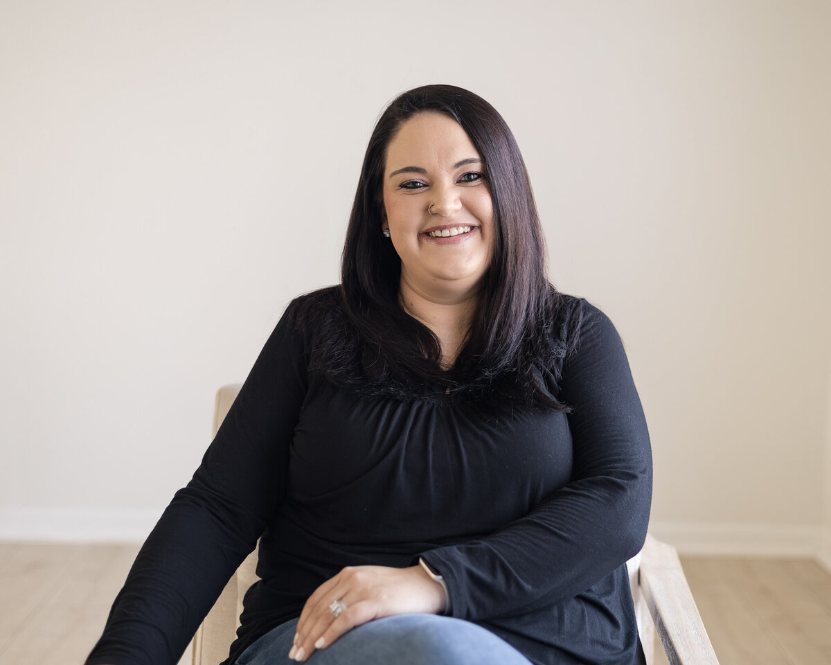 Woman sitting in a chair with legs crossed, has black hair, wearing a black shirt, blue jeans, and black shoes, smiling at the camera