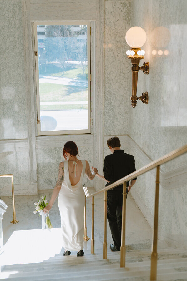 Holding hands, a man and woman walk down the stairs on the inside of the capitol building in Boise, ID. She's holding a wedding bouquet in her other hand.