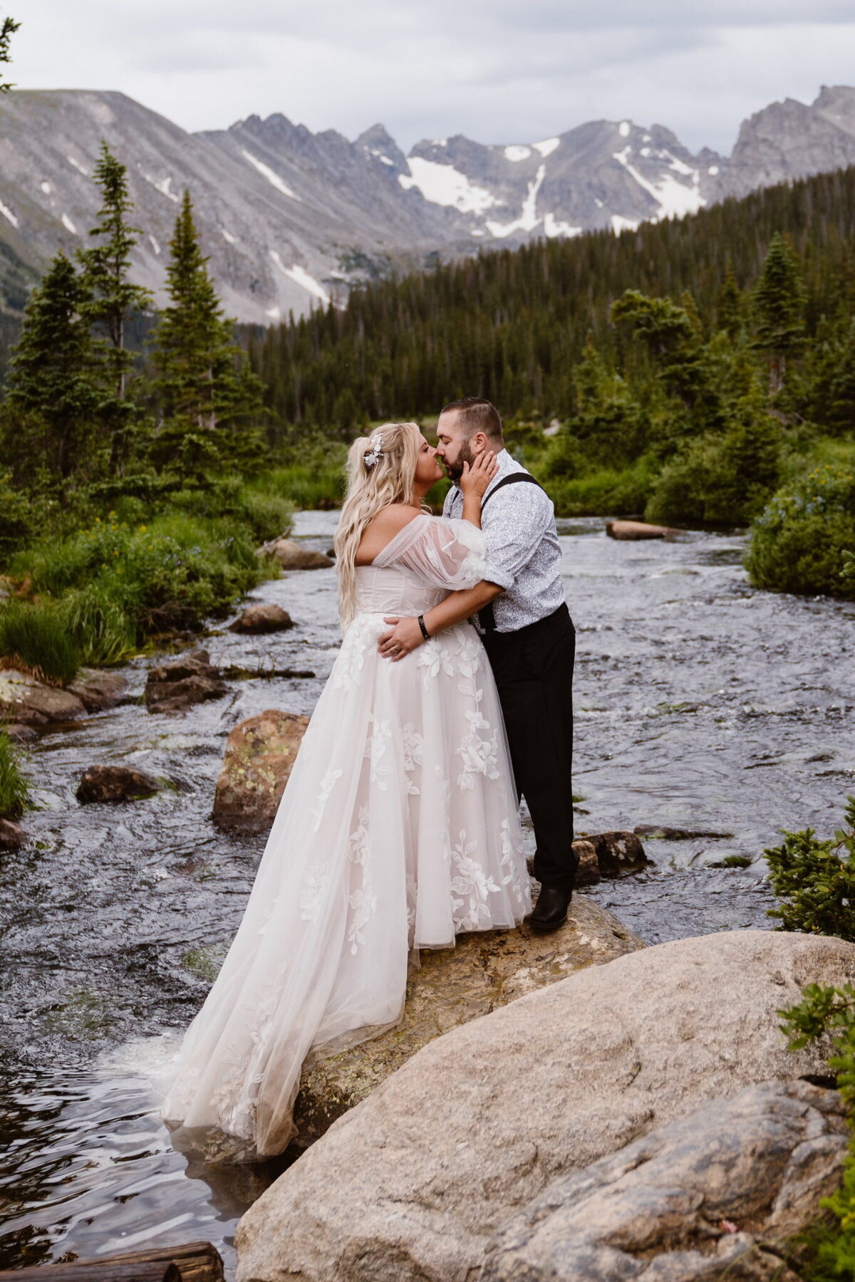 Bride and groom kissing on a rock in a mountain stream during their Brainard Lake Recreation Area, Colorado elopement, with dramatic peaks and pine trees in the background.