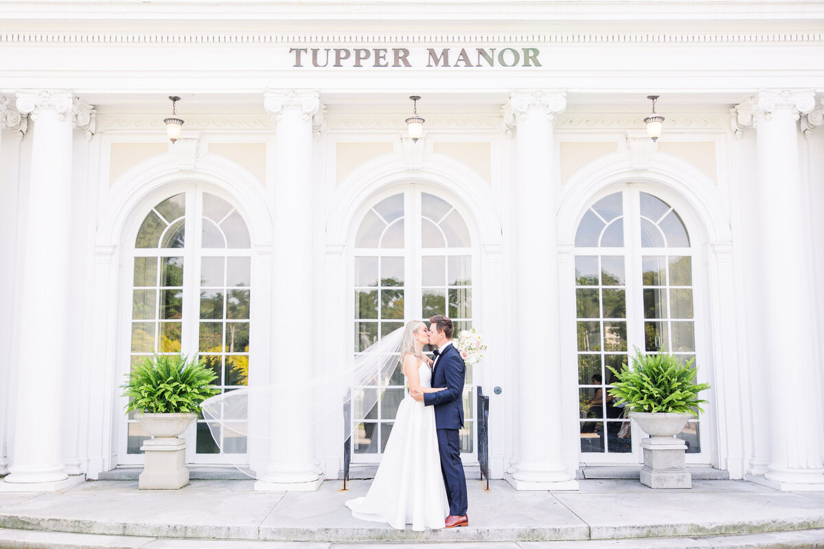 Bride and groom kissing with her veil blowing in the breeze in front of Tupper Manor