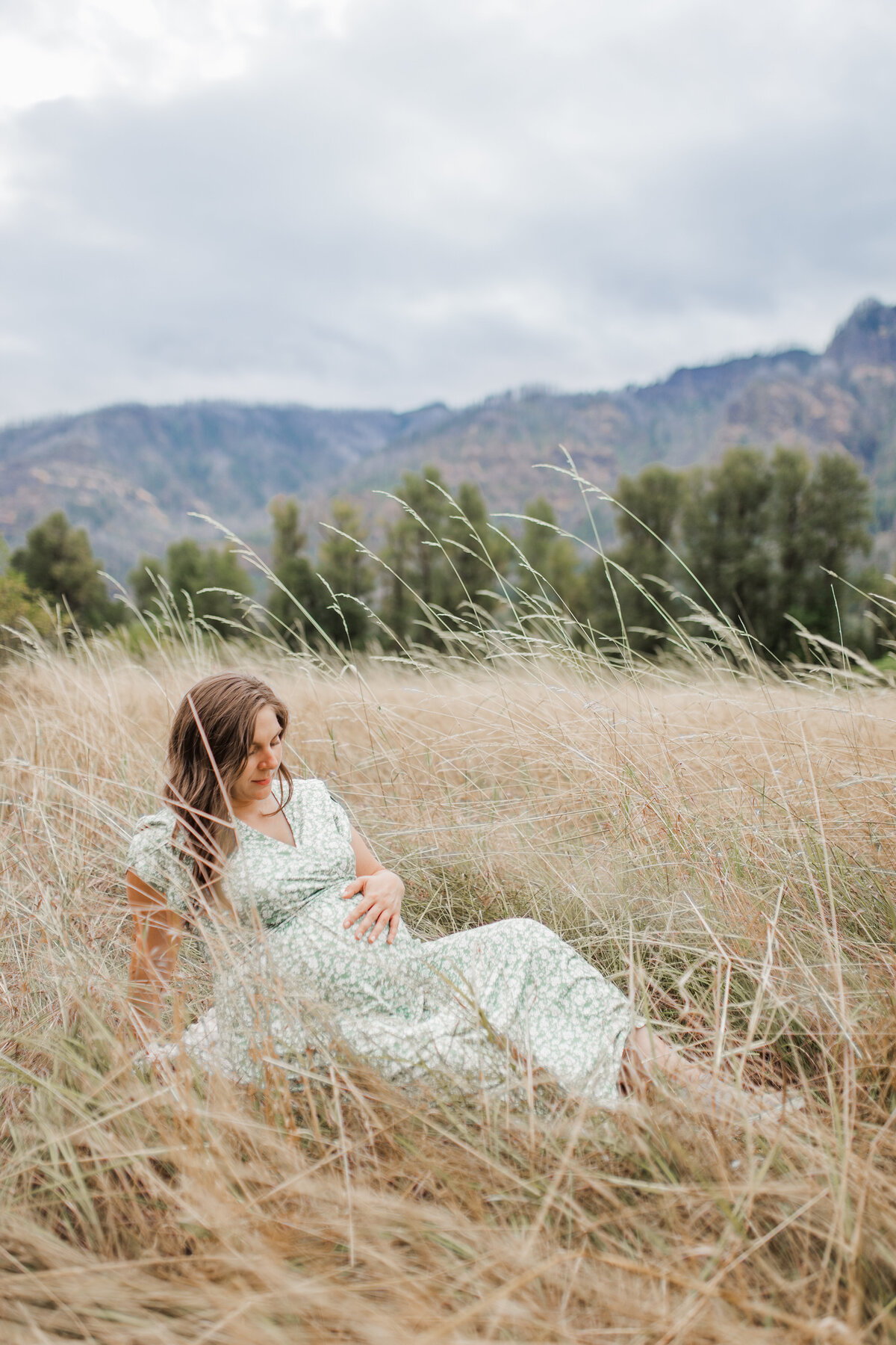 Pregnant woman sitting in tall grass with mountains in the background