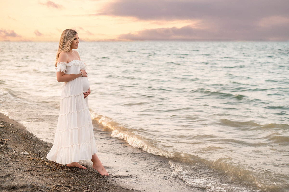 Pregnant mom at her Toronto portrait outdoor session standing at the edge of the water at the beach during sunset.