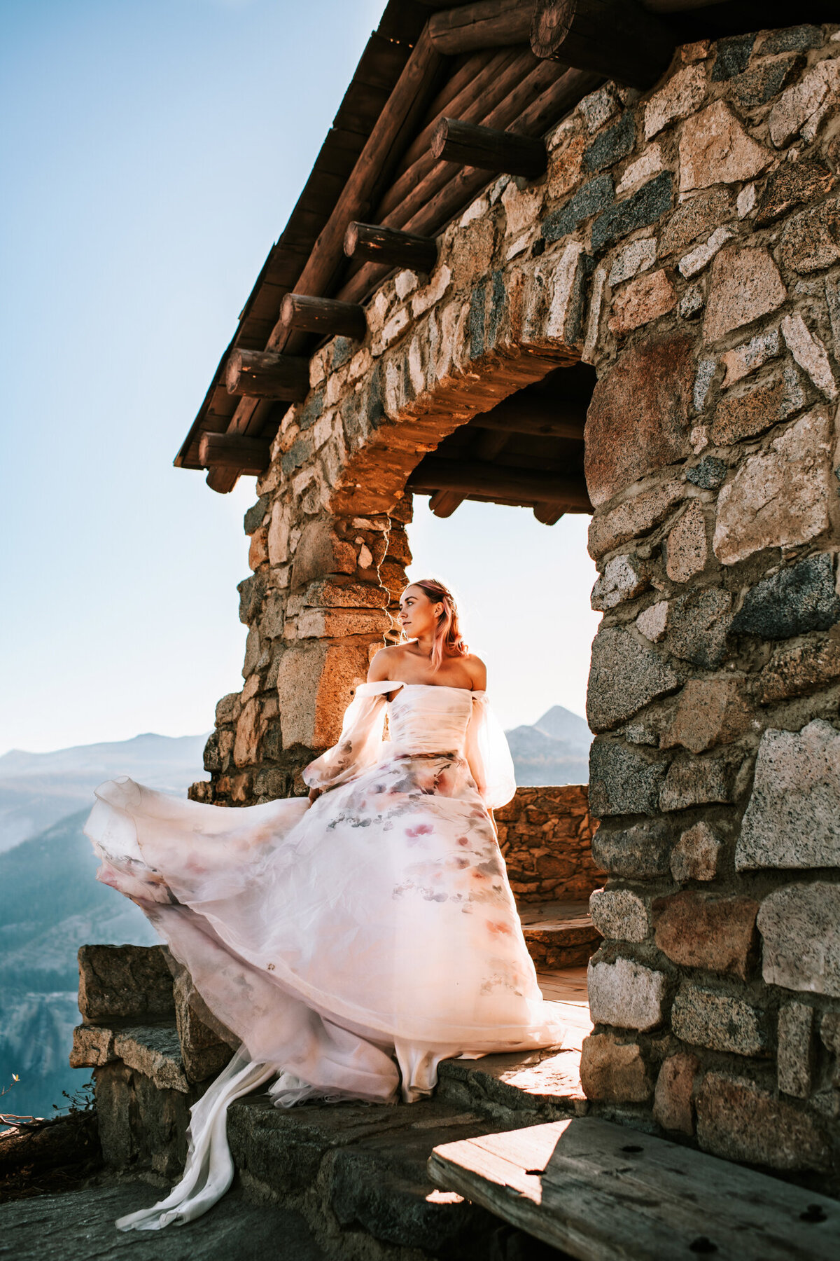 Couples Photography, Woman in a wedding dress stands in a stone covered awning on top of a cliff at Yosmite, looking out into the sun.