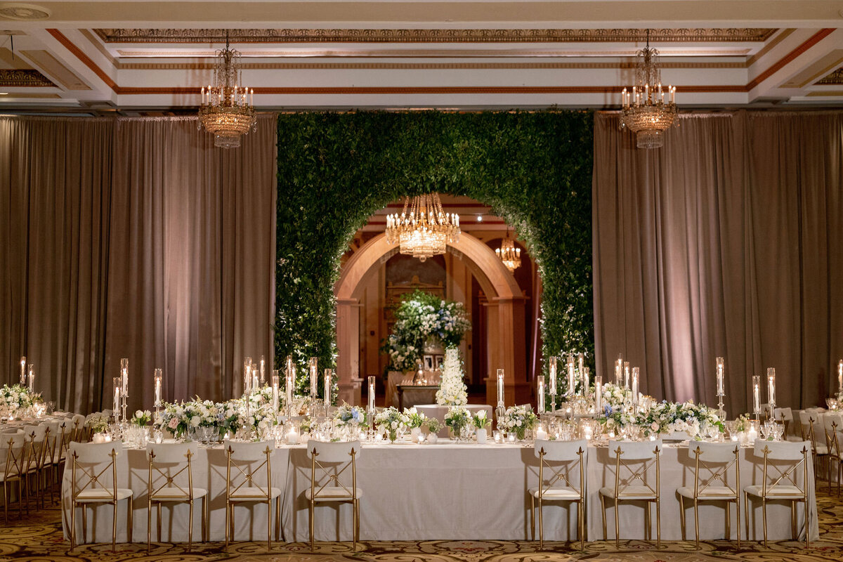 Elegant indoor wedding reception with tables set for dinner and a large floral arch at the front of the hall.