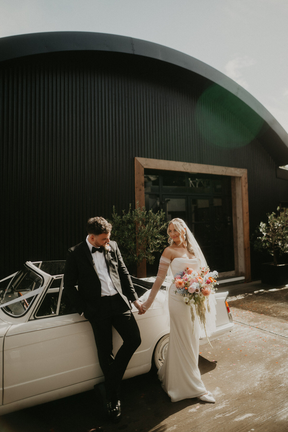 Bride and Groom stand in front of a vintage Triumph with the top down at The Shack Revolution on their wedding day. 