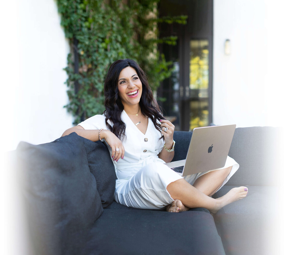 A professional and candid brand photo of Stefanie Gass smiling and sitting cross legged on an outdoor sectional with her laptop open on her lap