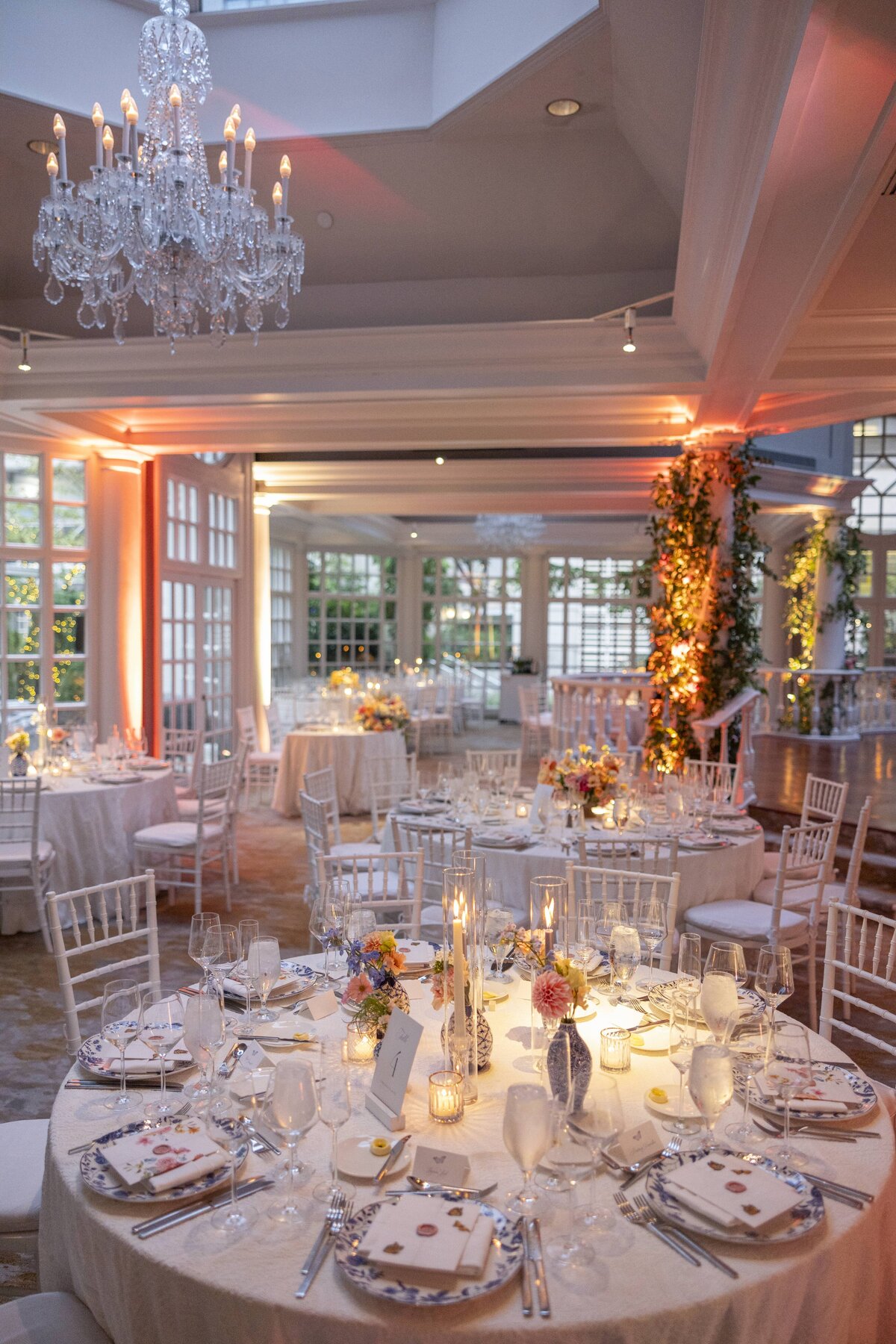 Elegant wedding reception with round tables set with white tablecloths, floral centerpieces, and tableware. A large chandelier hangs from the ceiling, and the room is lit warmly with natural light streaming through large windows.