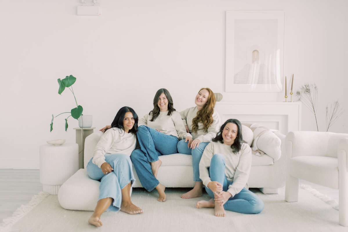 Team of women in matching crewneck sweaters and jeans, barefoot on cream sofa—candid brand unity
