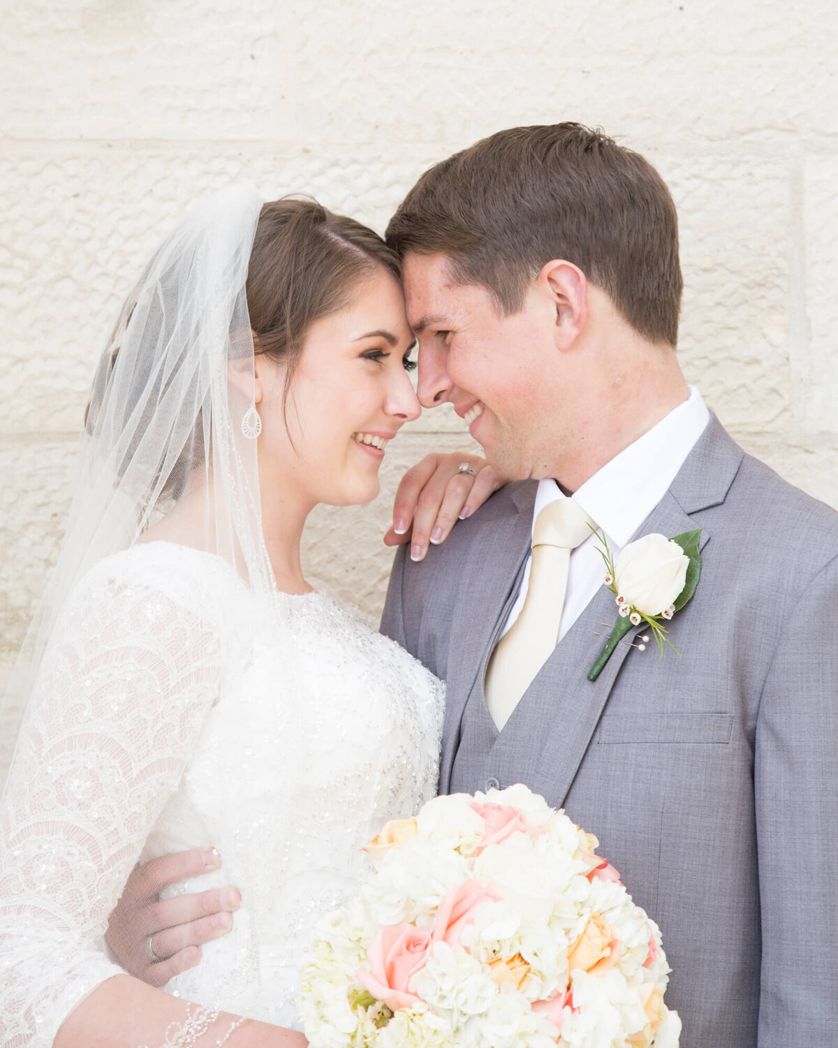 groom in gray suit holding bride in lace and beaded dress with a veil against a stone wall at beautiful lds temple wedding