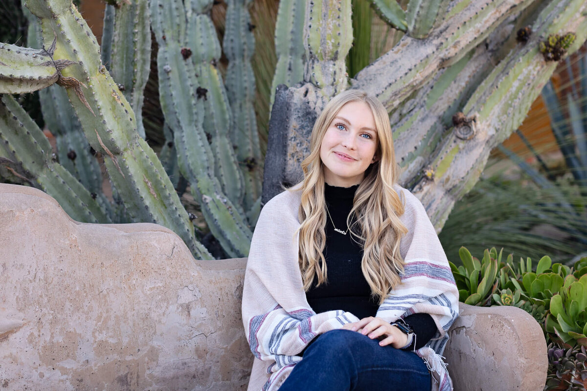 A young woman with long, blonde hair in blue jeans, black turtleneck, and beige cardigan, sitting on a clay bench with a cactus behind her, smiling softly at the camera.