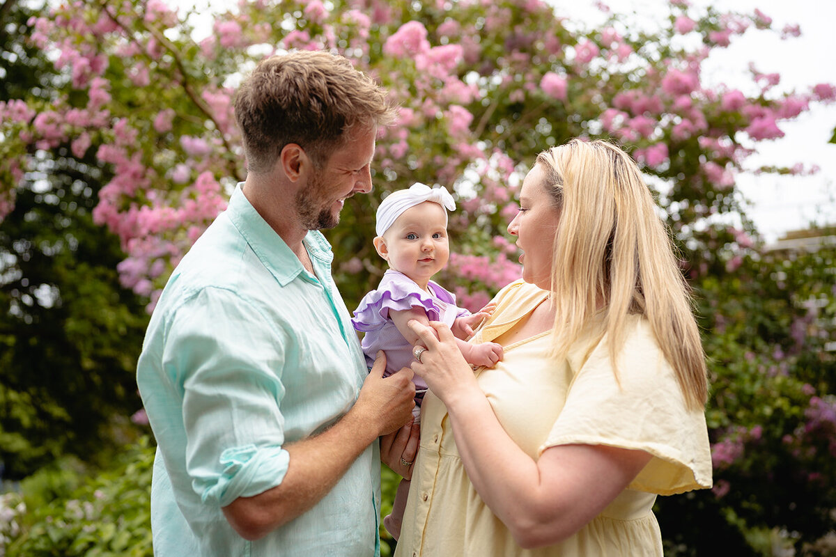 Young family portrait in Geelong featuring parents holding their baby girl in a garden with blooming pink flowers.