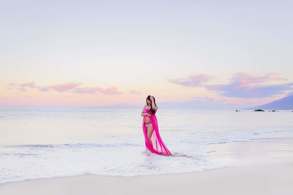 Woman in bright pink maternity gown poses on the beach at sunrise in Wailea while being photographed by Love + Water
