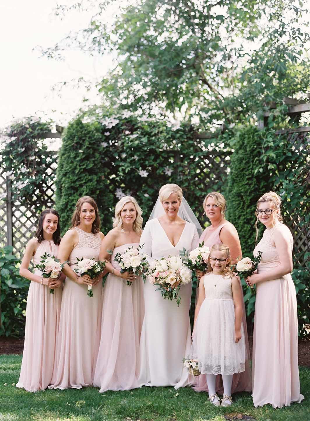 bride and brides maids in blush pink dresses in garden with greenery trellis