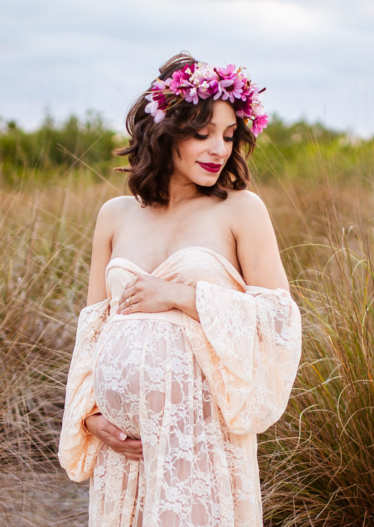 Pregnant woman hugging pregnant belly with pink floral crown while standing in tall grass