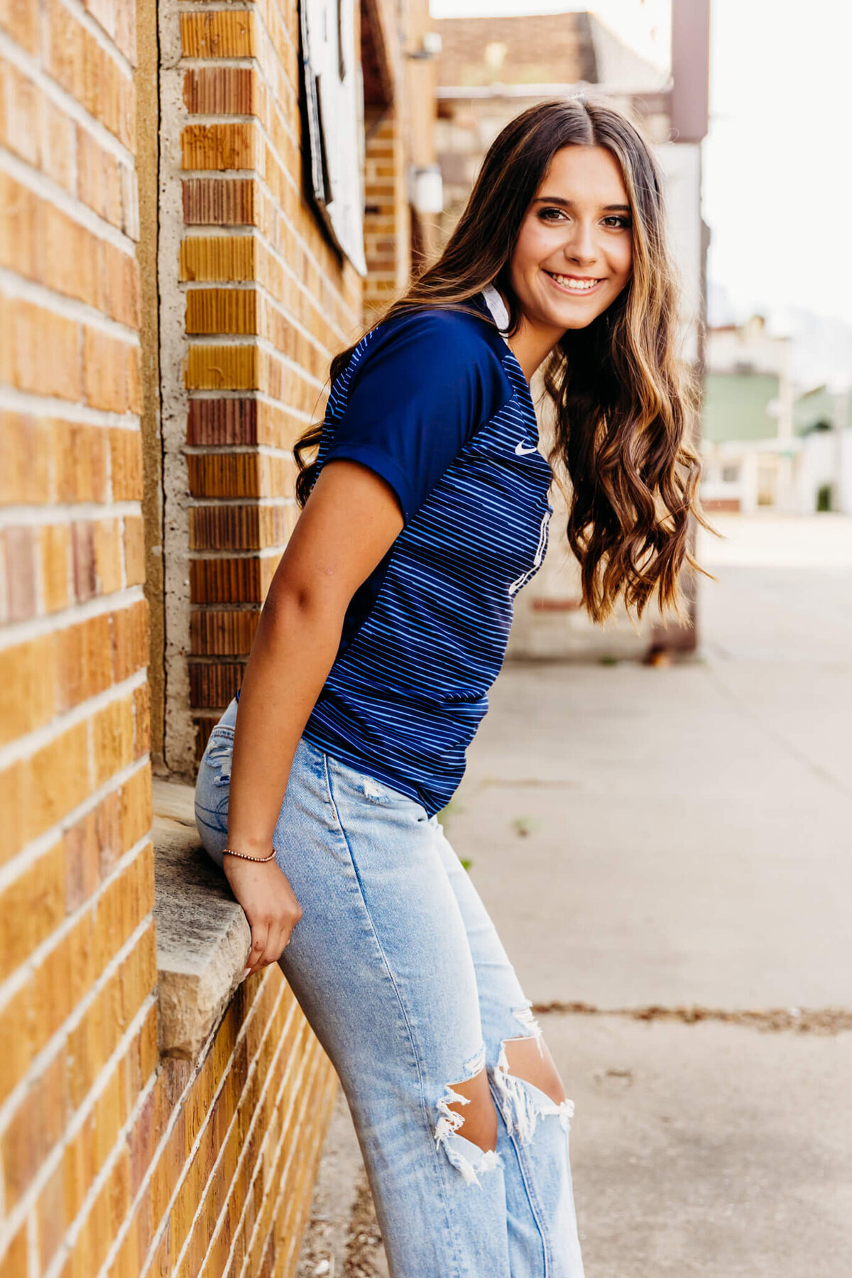 gorgeous teenage girl sitting on a window sill in front of a brick building as she leans forward and looks over her shoulder captured by Ashley Kalbus Photography