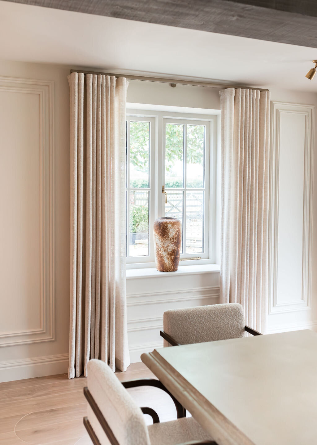 A large window with light colored curtains behind a modern dining table. The window has a large cream  and brown vase on the windosill.