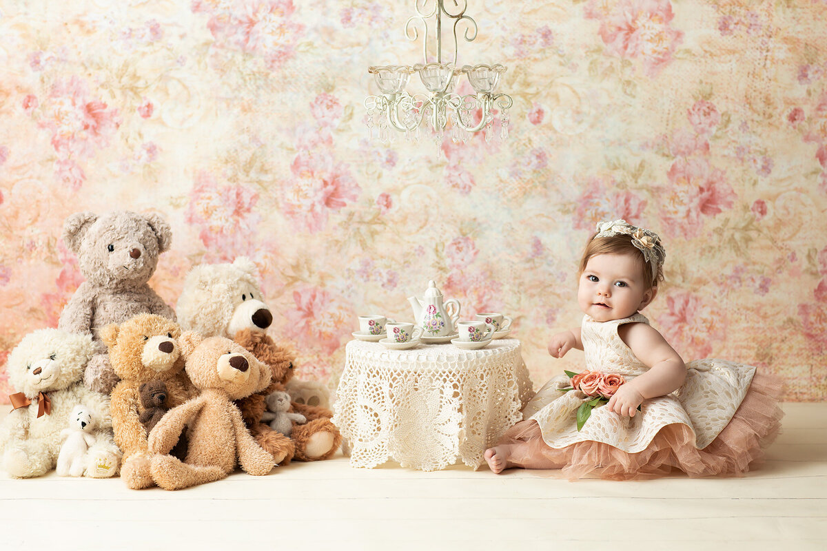 Baby girl in a cream and peach dress having a tea party with stuffed animals under a vintage chandelier, styled by a Dallas milestone photographer.