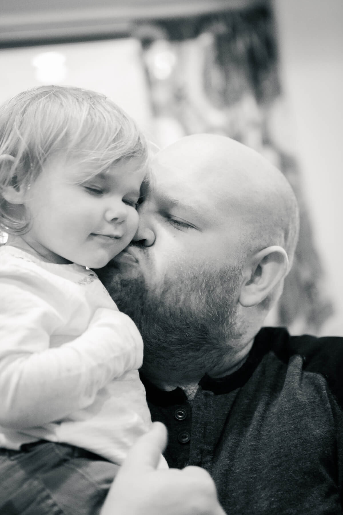 A toddler snuggles up to her daddy's cheek as he leans in to give her a kiss on the cheek.