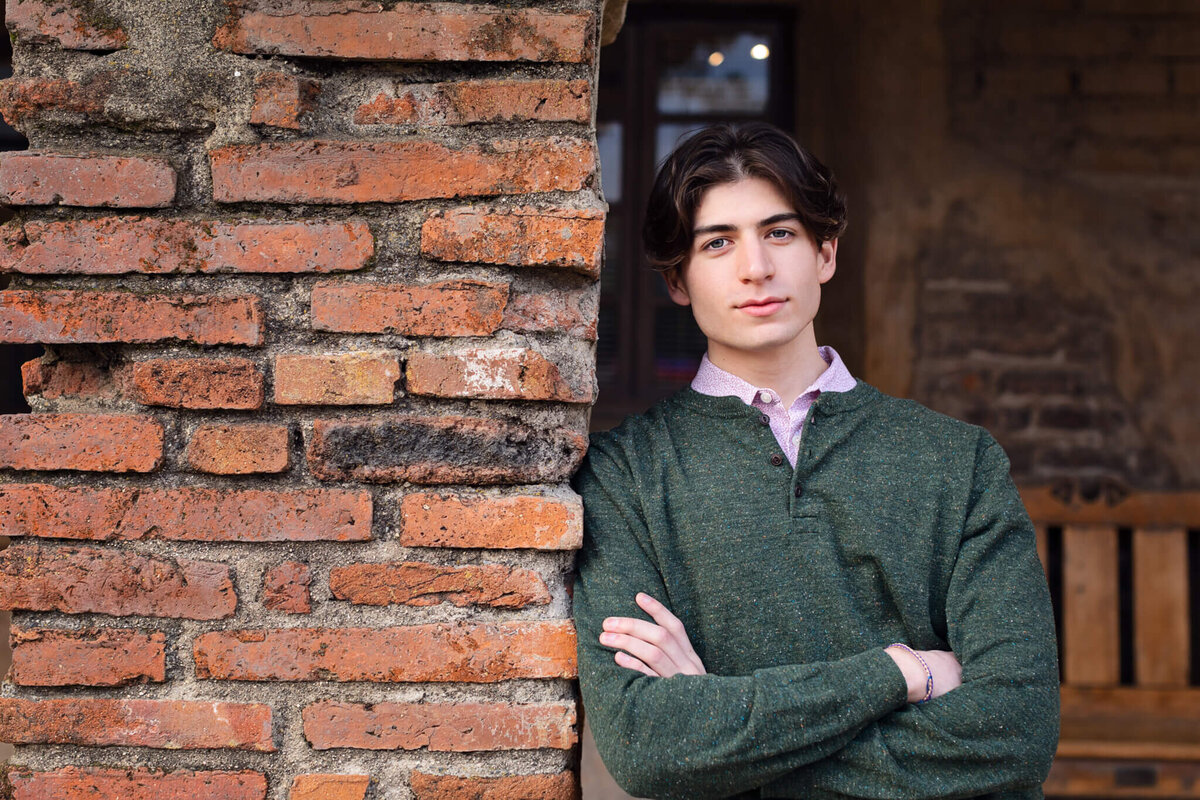 A young man with black hair wearing a green, long sleeve shirt crossing his arms and leaning against a brick wall, looking seriously at the camera.