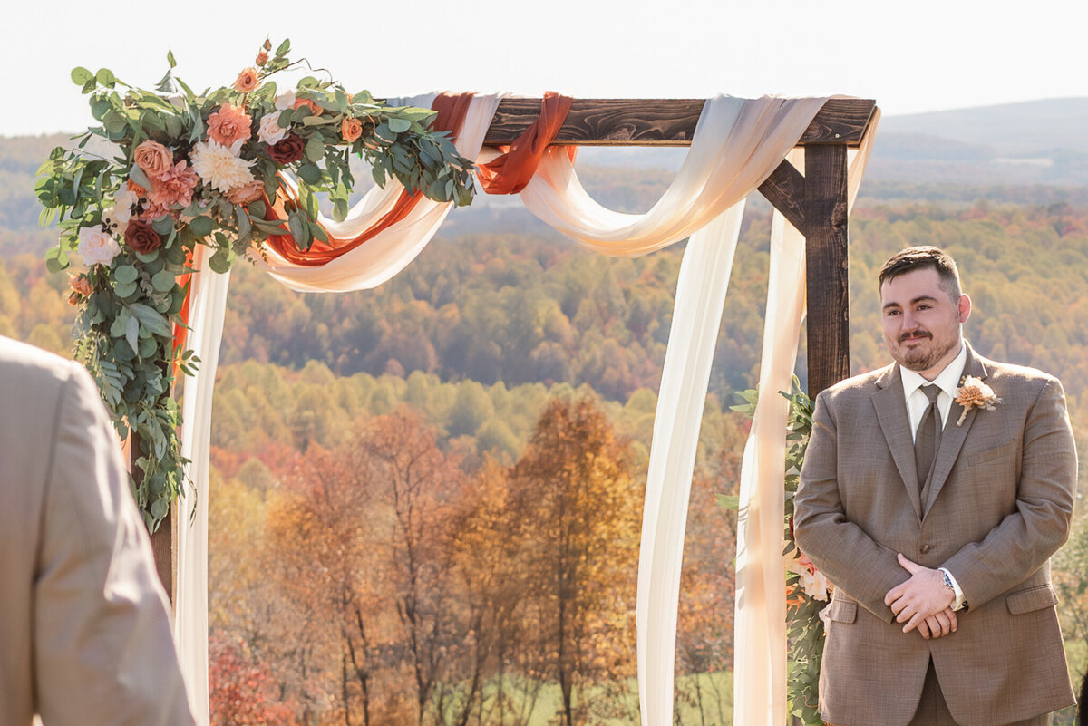 Groom smiles as he sees his bride walking down the aisle during a fall wedding ceremony in the Pennsylvania mountains -  captured by Bree Stewart of Lens Love Photography.