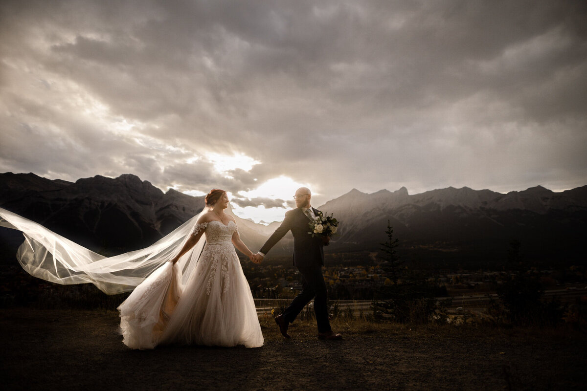 Gorgeous moody portrait of bride and groom walking holding hands in the mountains captured by TkShotz, modern wedding photographer and videographer in Calgary, Alberta. Featured on the Bronte Bride Vendor Guide.