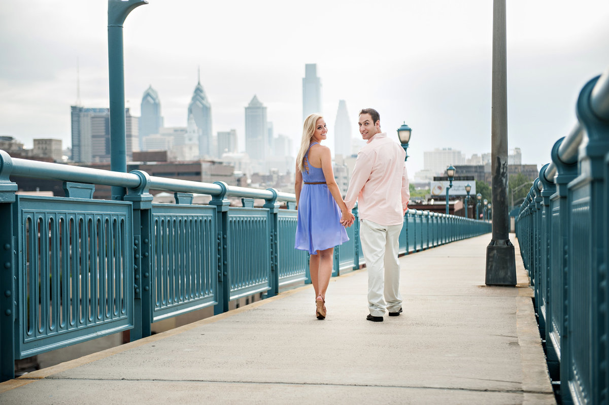 A couple walk on the ben franklin bridge with the philadelphia skyline behind them.