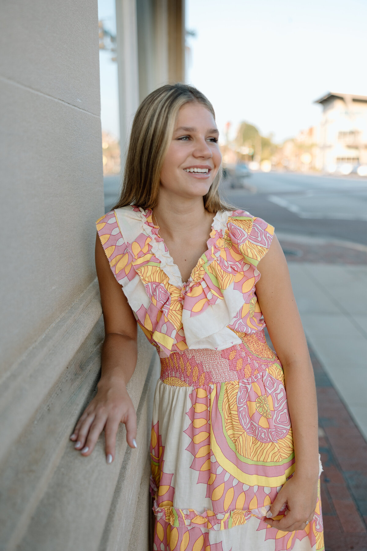 Girl wearing long colorful sundress in Downtown Sumter, SC leans against a wall and smiles