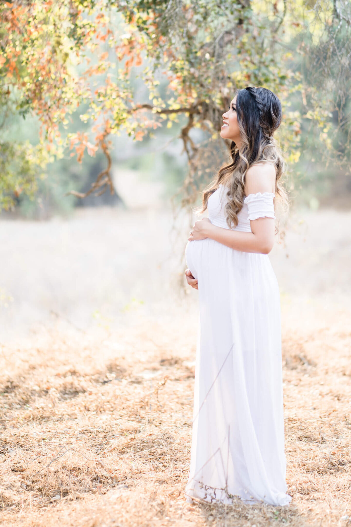 Pregnant mom holding pregnant belly under autumn tree leaves with crown braid in her hair and loose waves. Maternity inspo photo by Orange County Maternity Photographer Melliemade Photography