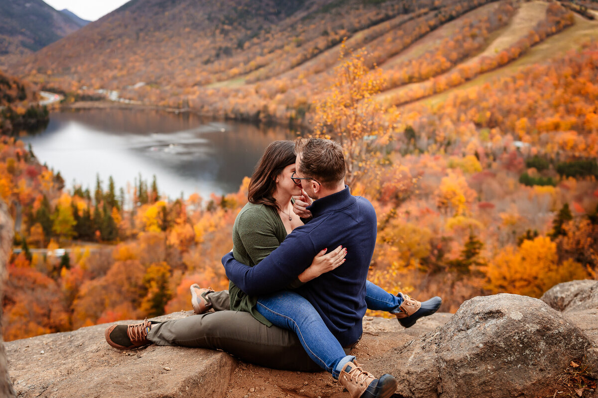 Engaged couple sitting on rocks at Artist bluff looking over White Mountain valley and Echo Lake in Lincoln NH