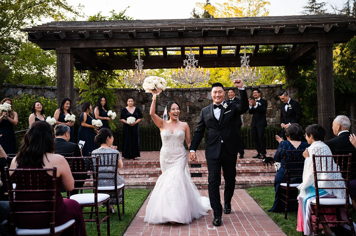 San Francisco Bay Area wedding photographer captures joyful recessional as bride and groom celebrate with raised bouquet under chandelier pergola at Estate Yountville in Napa Valley