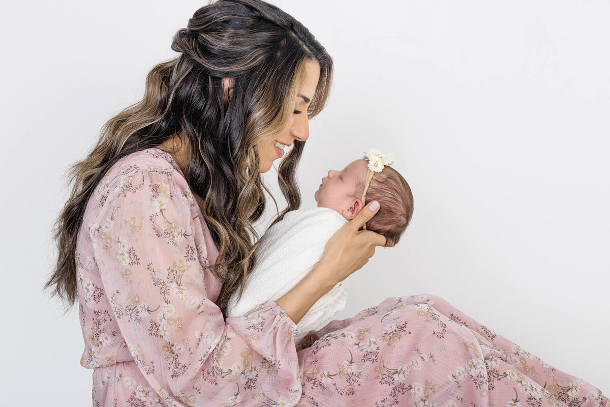 A woman with long, wavy hair, wearing a floral pink dress, lovingly cradles a newborn dressed in white. The baby has a delicate headband with a flower. They sit against a plain white background, sharing a tender moment.