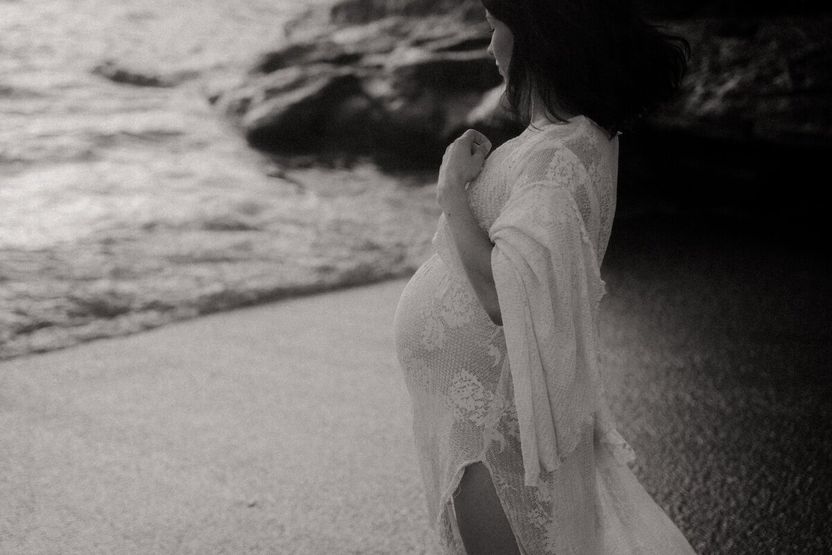 La future maman en dentelle blanche regarde l’océan. Noir et blanc, ambiance intime. Photo réalisée par Kamescop, photographe maternité.