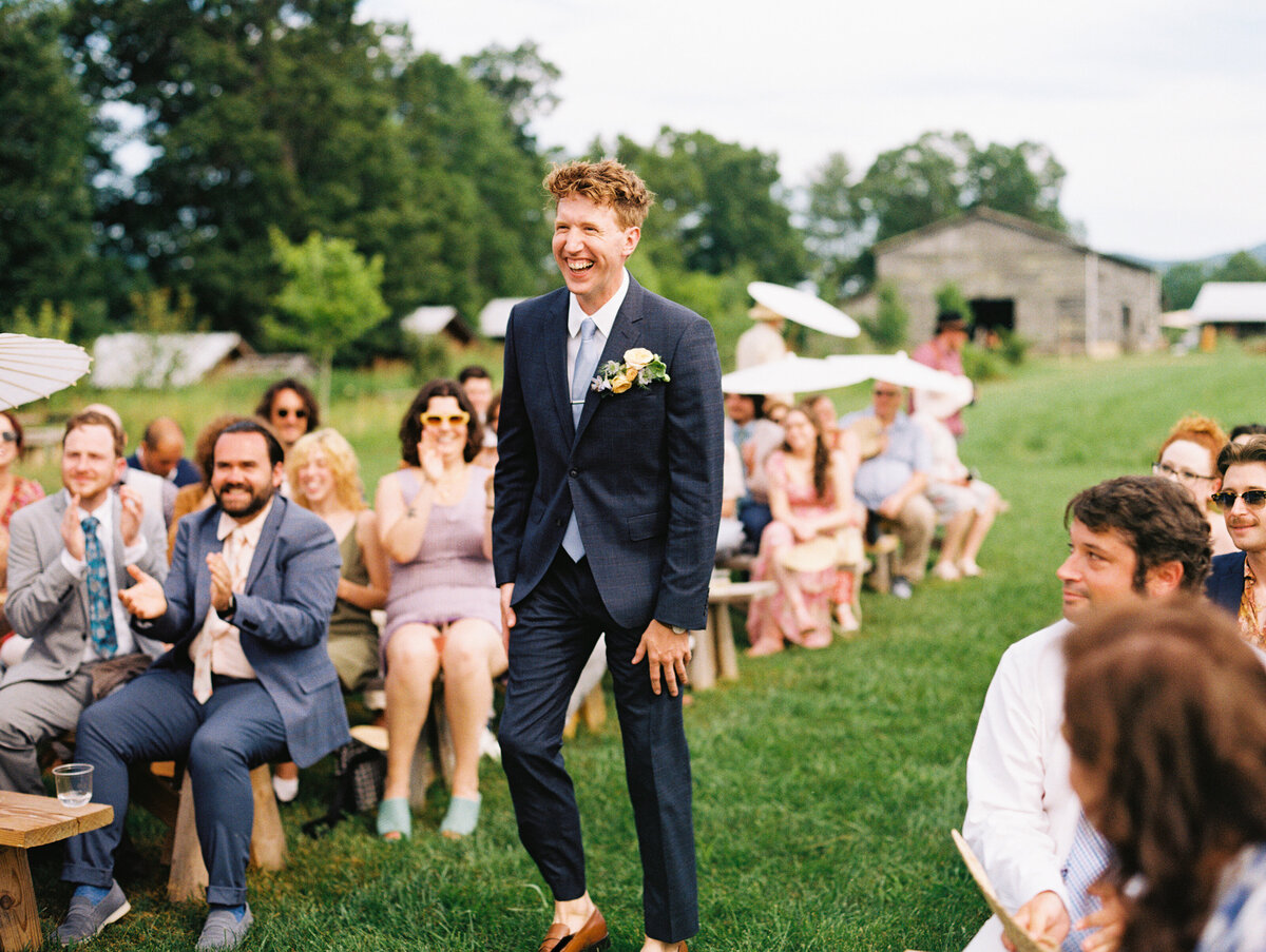 A beaming groom makes his entrance to the cheers of wedding guests, wearing a boutonniere by Moonlight Floral Co., with the barn and cabins in the background at Paint Rock Farm in North Carolina, by film photographer Megan Lynn of My Sun and Stars Co.