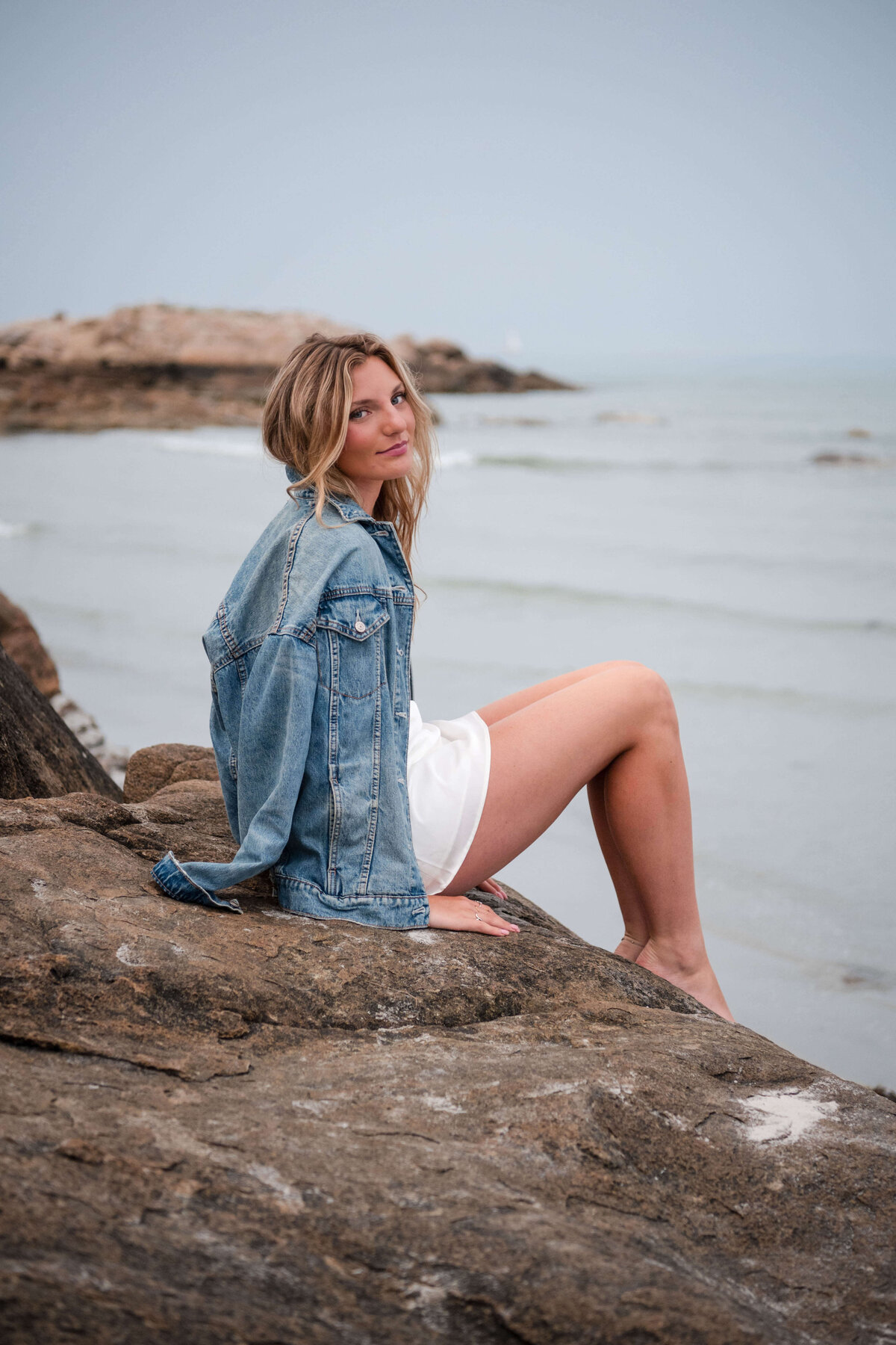 A senior sits on a rock and smiles back at the camera with a jean jacket draped over her and Wingaersheek Beach in the background.