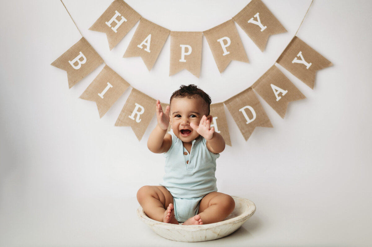 One year old baby boy wearing baby blue romper sitting inside of a wooden round bowl in front of a  white backdrop with a tan happy birthday sign