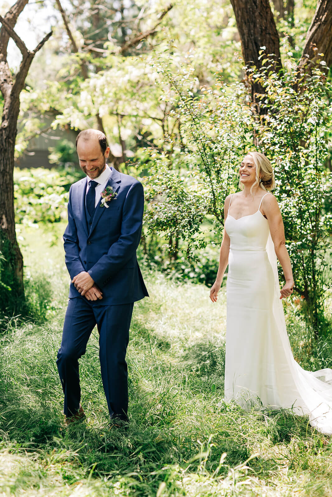 A groom stands outdoors with his back to the bride.