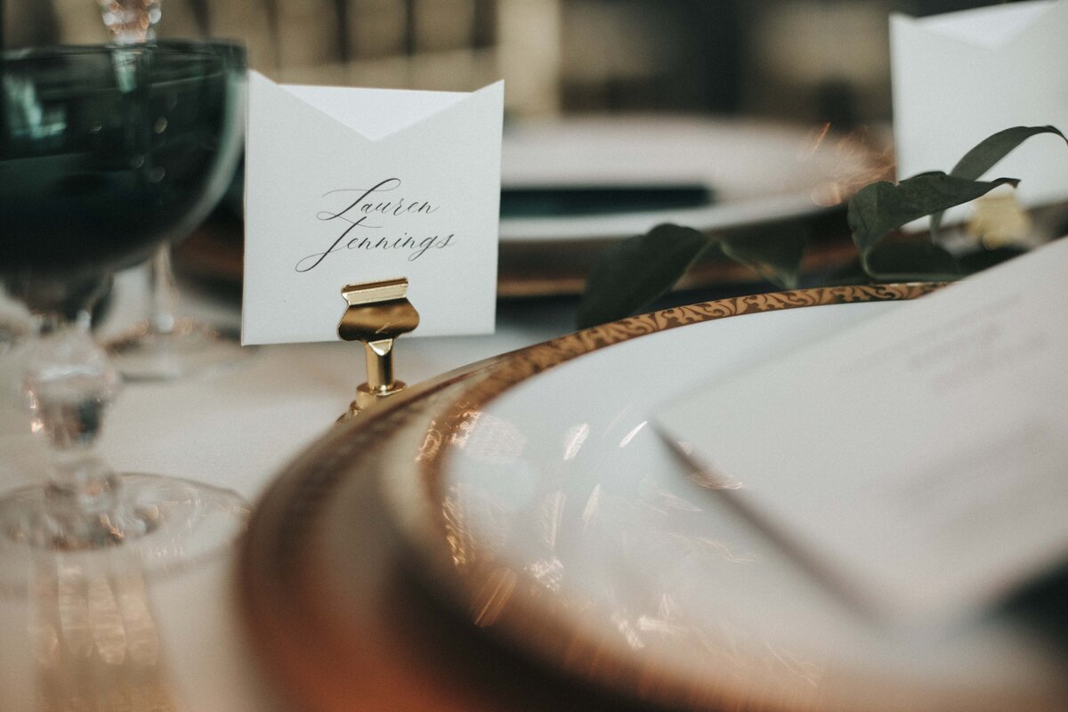 White square place card with black cursive font on a gold place card holder set on a table with elegant plates and glassware.