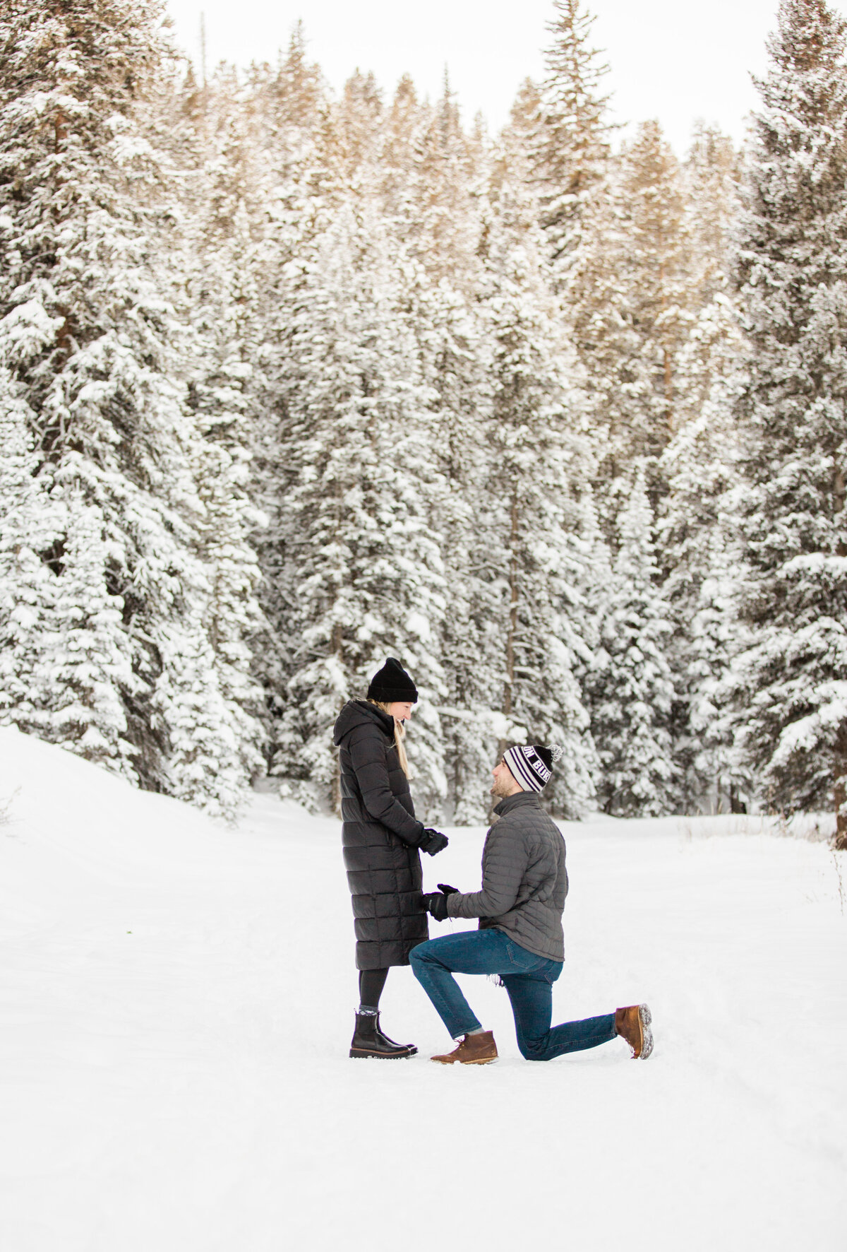 A man is down on one knee proposing to this girlfriend amidst a snowy winter scene