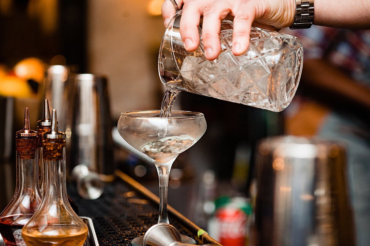 A bartender uses a cut crystal mixer to pour a signature wedding cocktail into a frosted coupe glass. Bottles of bitters sit on the bar beside the glass.