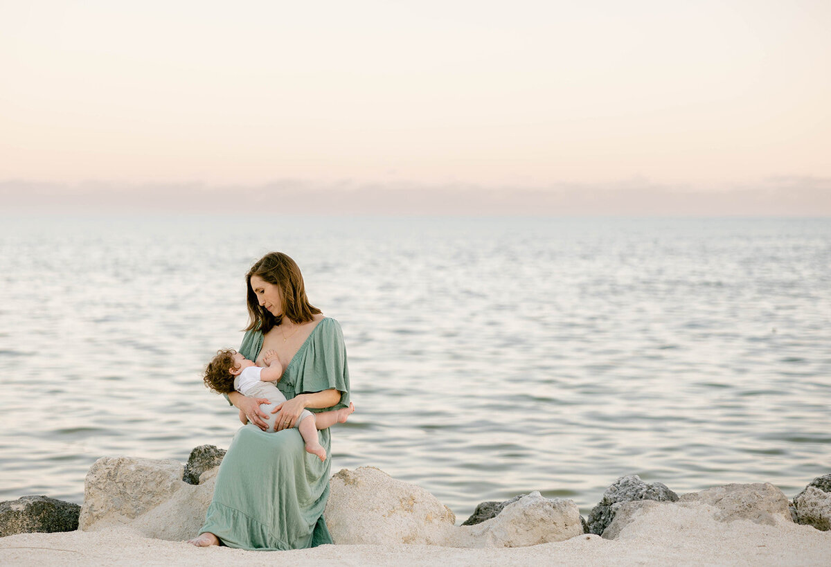 A mother in a flowing green dress holds her baby by the ocean during a Florida Keys family session. Claudia Amalia Family Photography