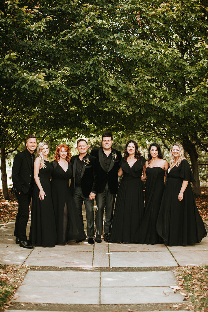 Two grooms wearing black tuxedos pose outdoors with their wedding party also wearing all black.