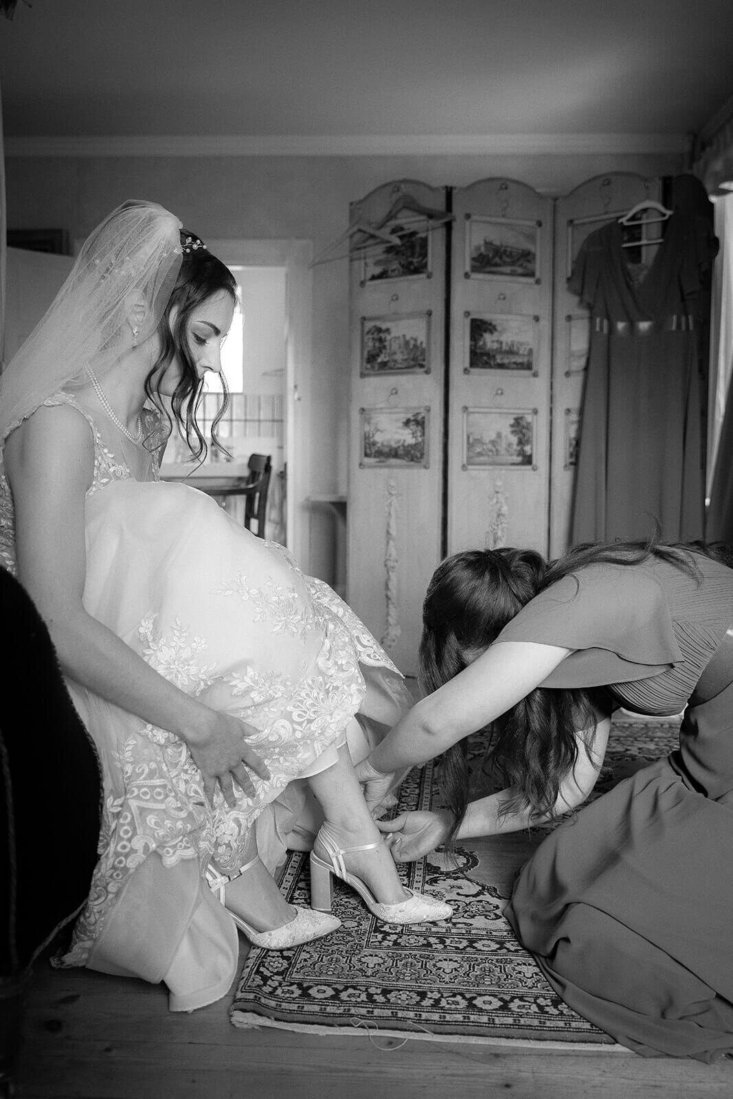 A maid of honour puts the brides shoes on her as she is seated