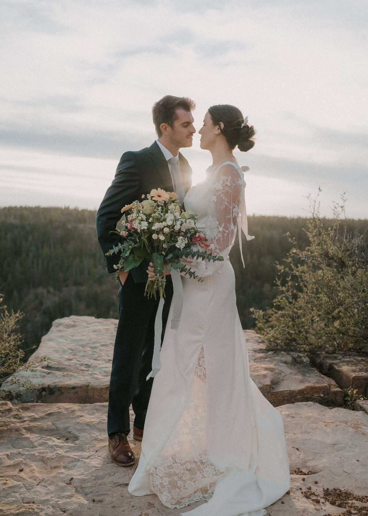 Bride holding bouquet and kissing groom with scenic view of Mogollon Rim in Payson, Arizona with sun shinning between them