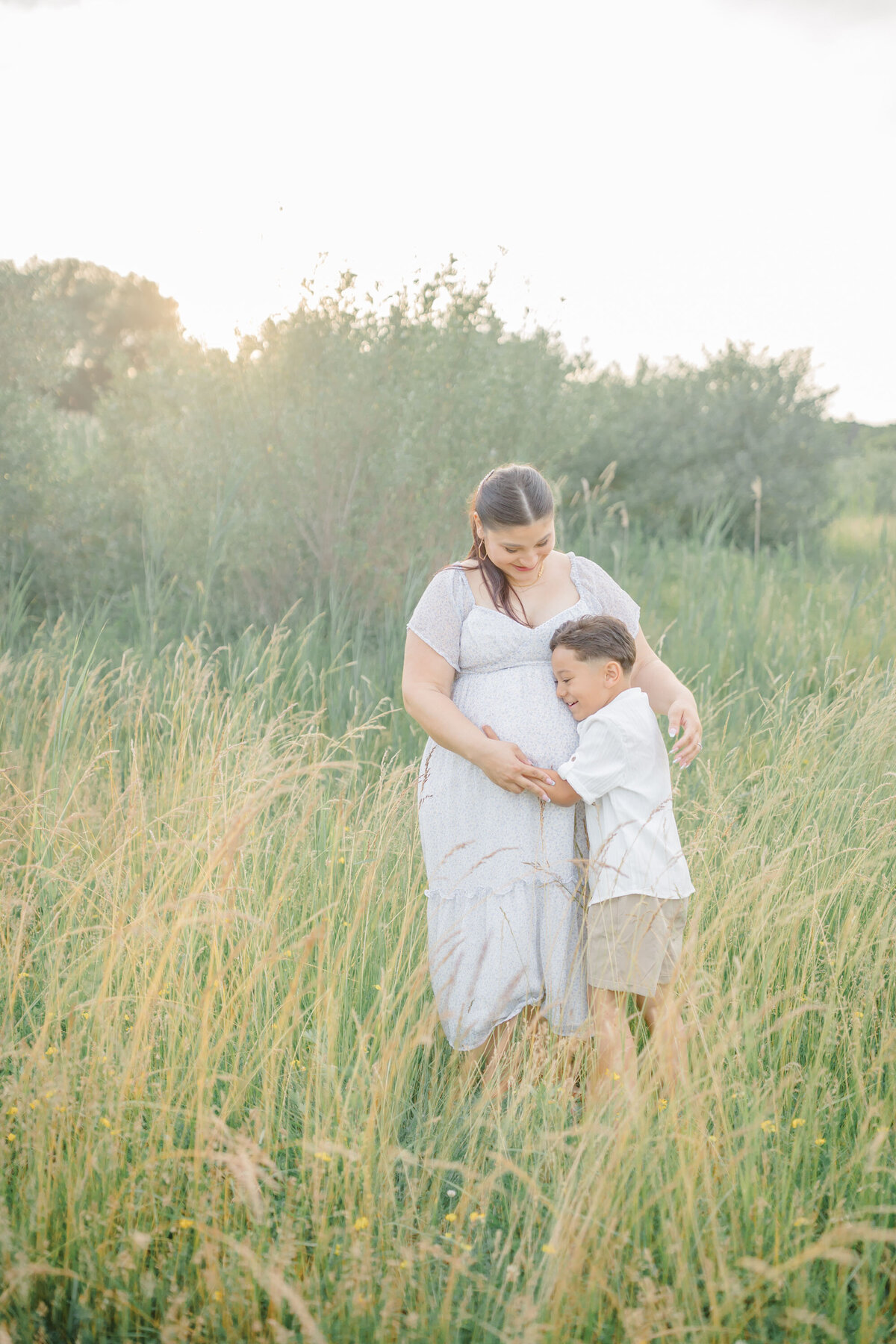 A mother stands in a serene outdoor setting, her young son gently hugging her pregnant belly. She smiles warmly as he rests his head against her, embracing the growing bond with his soon-to-arrive sibling. Soft natural light filters through the trees, creating a tender and heartfelt moment between mother and son.