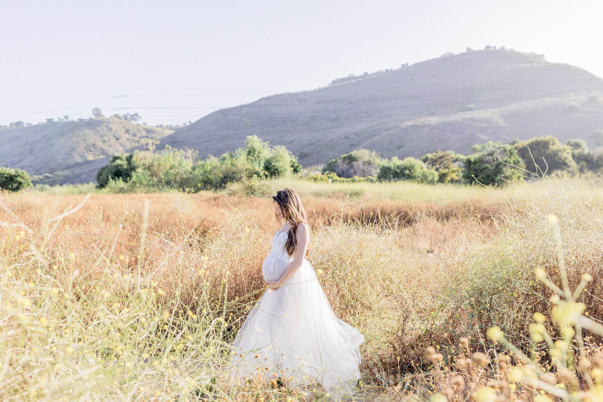 A pregnant woman in a flowing white dress stands in a sunlit field of tall grasses, with rolling hills and sparse trees in the background, under a clear sky.