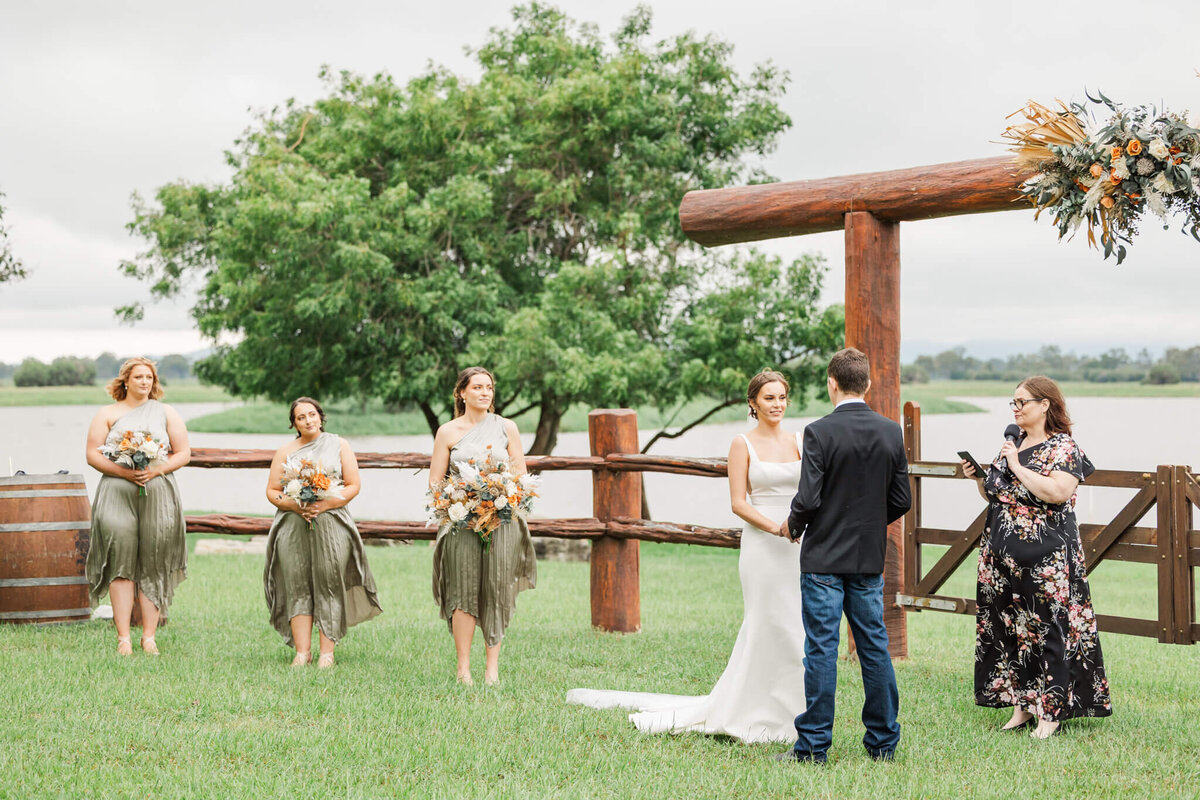 Bride and groom exchange vows in front of a wooden fence and gate at Paradise Lagoons, Rockhampton while bridesmaids watch from the side.