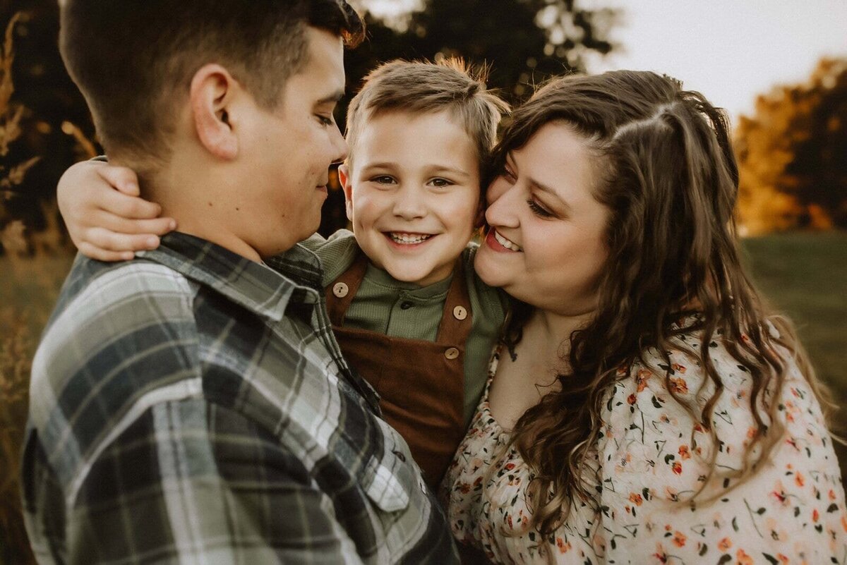 A couple is holding a smiling child between them outdoors. The man wears a plaid shirt, the woman a floral top, and the child a green outfit with brown overalls. Trees and grass are in the background.