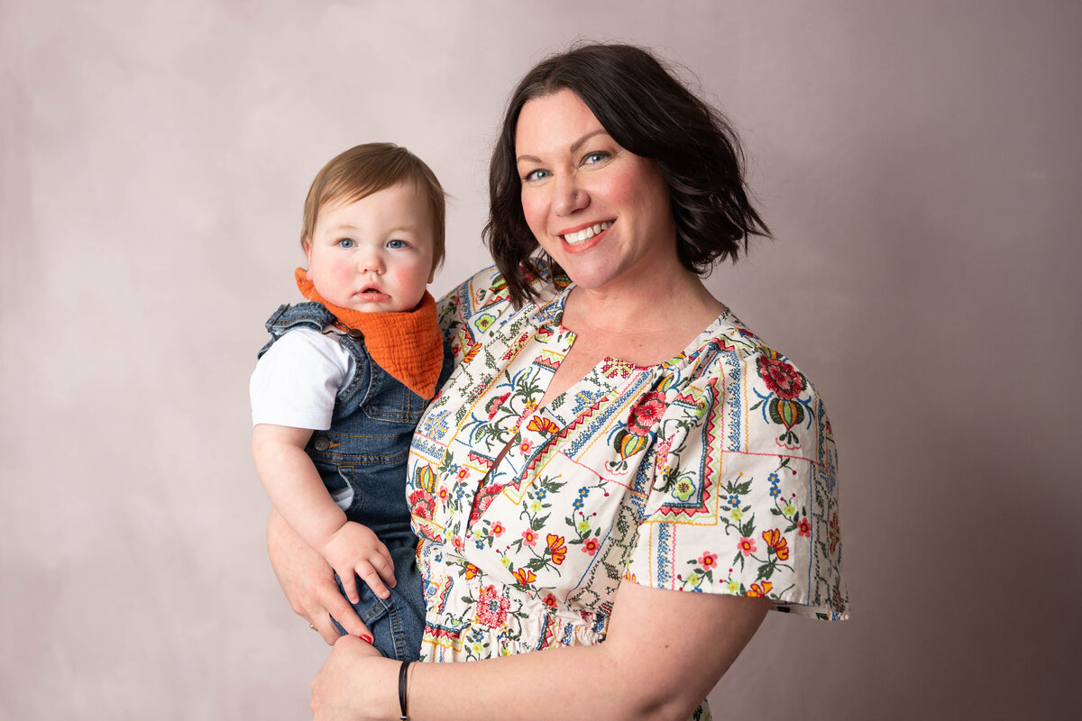 Baby wearing denim overalls being held by a woman in a tan dress with orange floral accents.